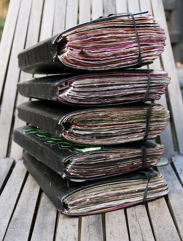 Une haute pile de carnets usés, tenus par des élastiques, sur un établi en bois