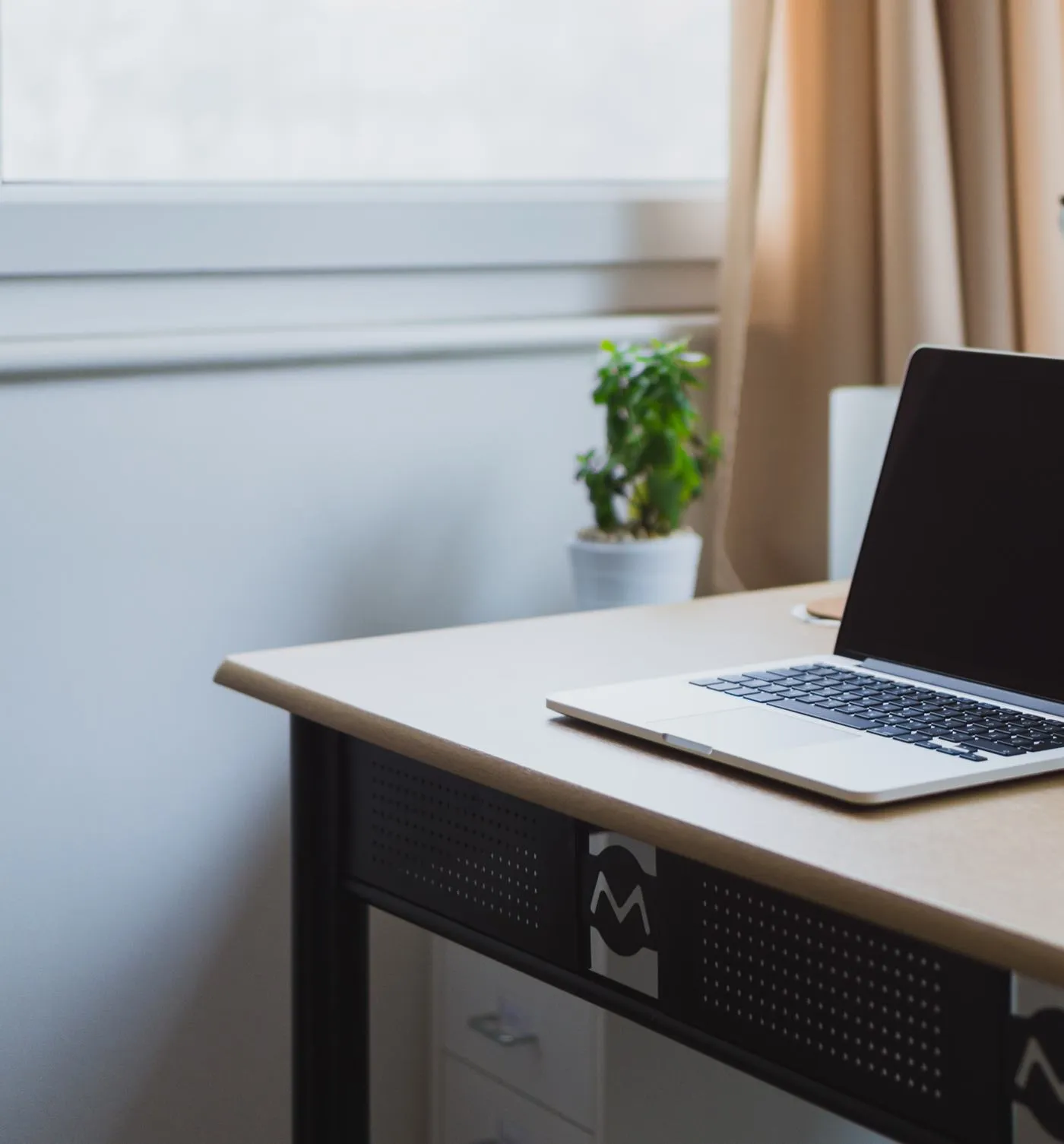 Un bureau clair près d’une fenêtre à rideaux, avec un ordinateur portable ouvert et une petite plante