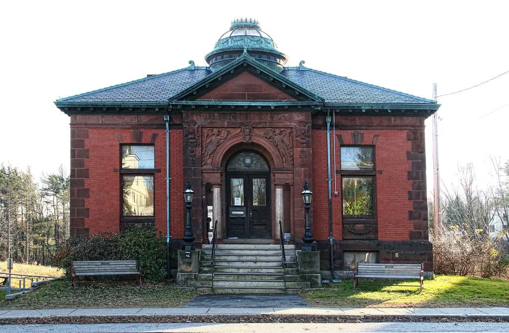 La façade en brique rouge d’une petite bibliothèque municipale, sa porte encadrée d’une arche et de deux lampadaires