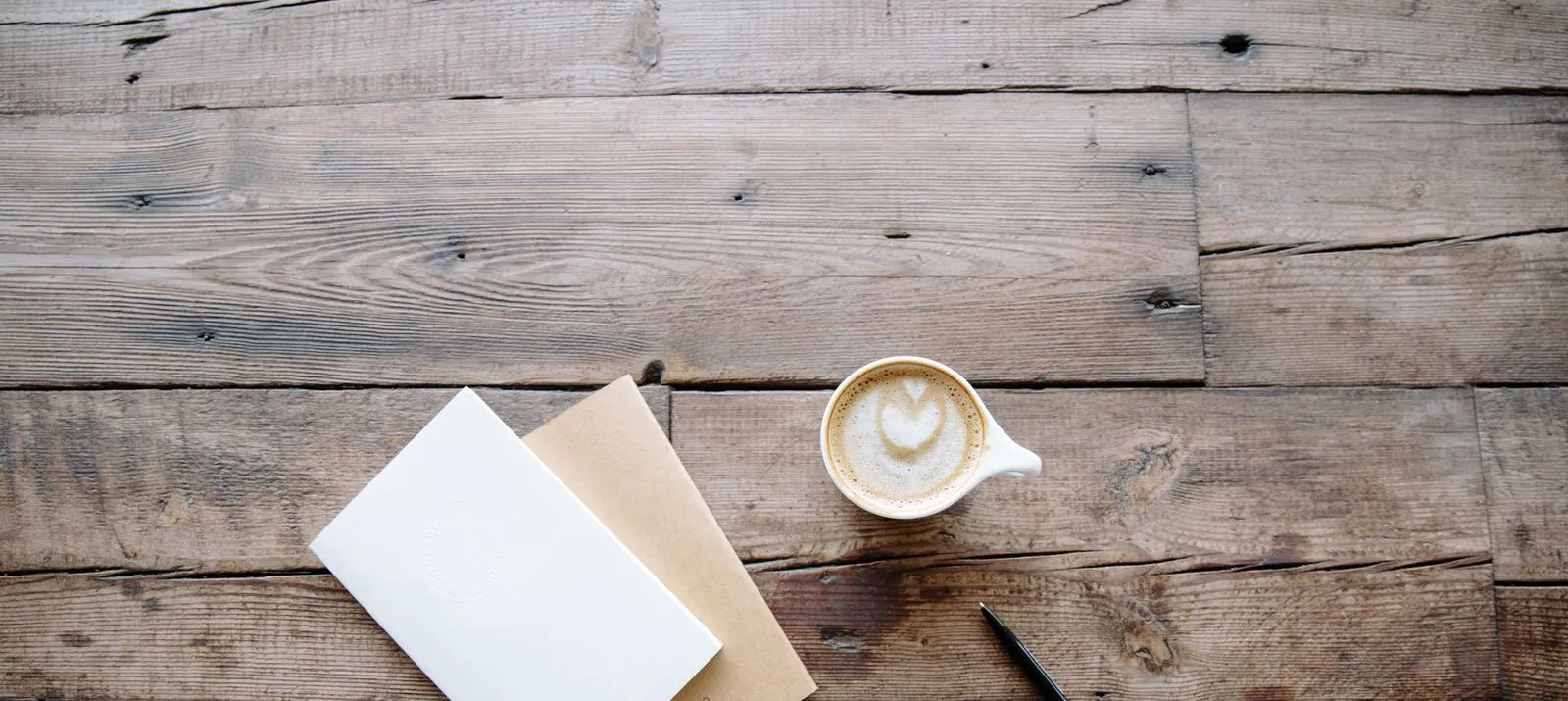 Un carnet fermé et une enveloppe kraft à côté d’un café au lait et d’un stylo sur une table en bois
